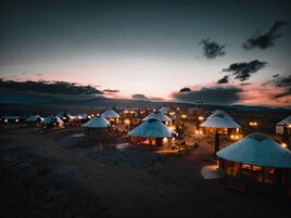 Diversas yurts com telhados cônicos, dispostas em círculo, iluminadas por luzes quentes sob um céu crepuscular.