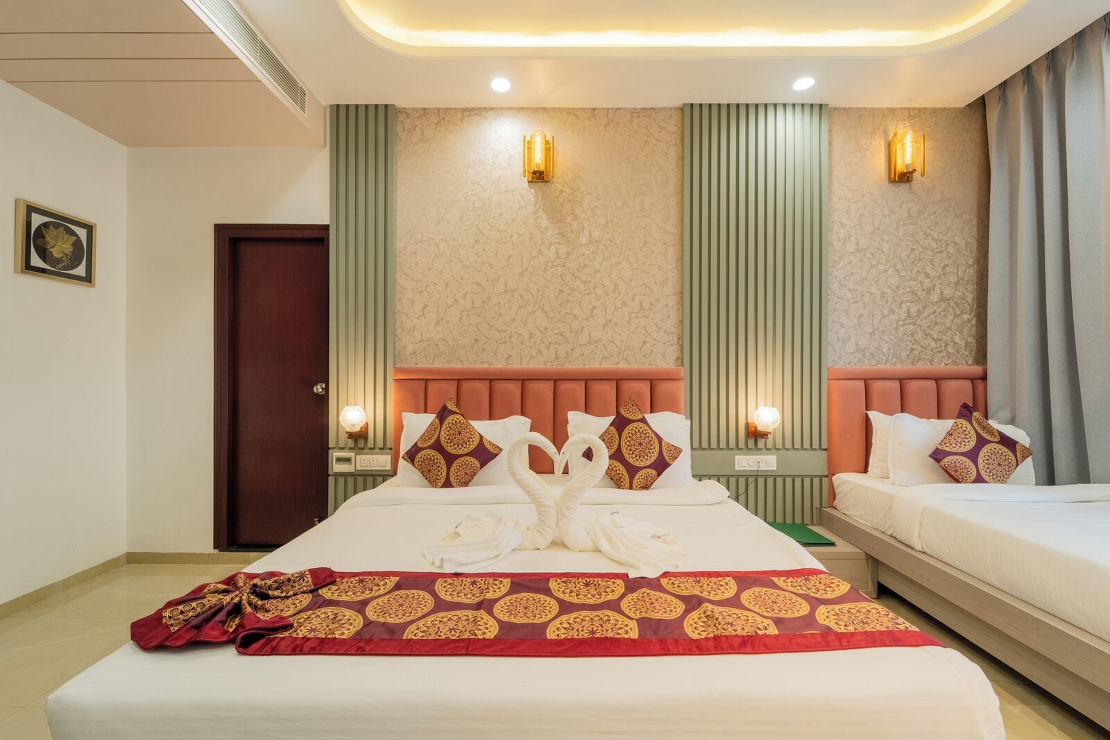 Une chambre d’hôtel avec deux lits, un couvre-lit rouge et doré à motifs, et un mur d’accent vert orné de lignes verticales.