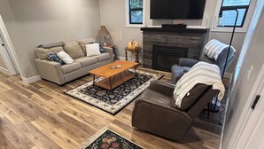 A modern living room with a fireplace, a sofa, a coffee table, and a TV. - Lake Breeze Cottage 6 beds w AC, hot tub near Lake Michigan beach and The Deck. (Muskegon)