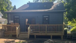 A two-story building with a dark exterior, a wooden deck, and a front door. - Lake Breeze Cottage 6 beds w AC, hot tub near Lake Michigan beach and The Deck. (Muskegon)