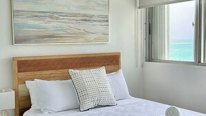 A bedroom with a wooden bed frame, white bedding, a ceiling fan, a painting of a beach, and a window with a view of the ocean. - Ocean View Apartment in Condado (San Juan)