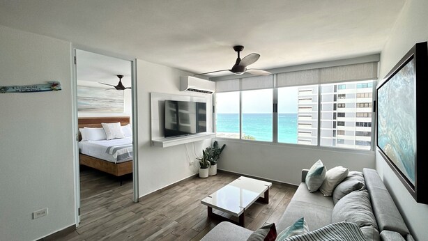 A modern hotel room with a bed, a TV, a coffee table, a sofa, and a view of the ocean. - Ocean View Apartment in Condado (San Juan)