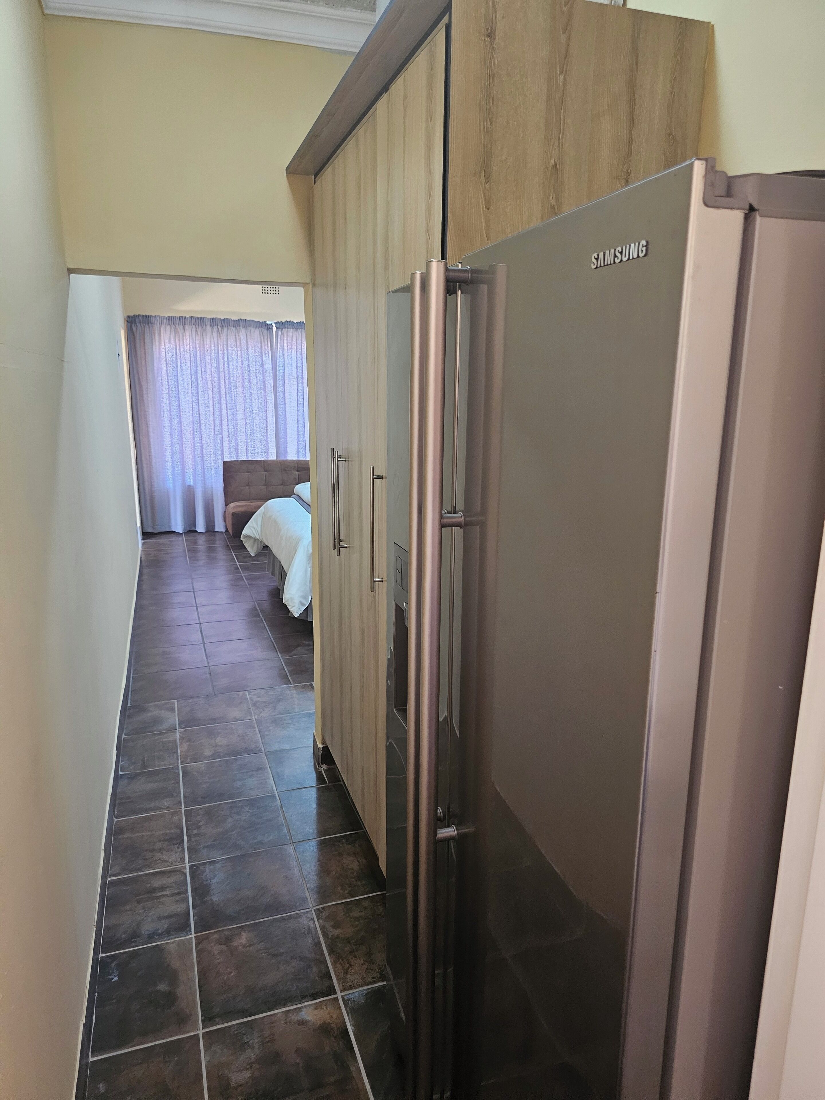 A corridor with a Samsung refrigerator, wooden cabinets, a bed with white bedding, and a curtain.
