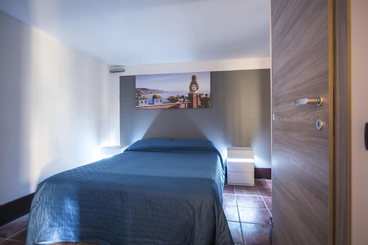 A bedroom with a bed, a nightstand, a wall-mounted shelf with decorative items, and a door.