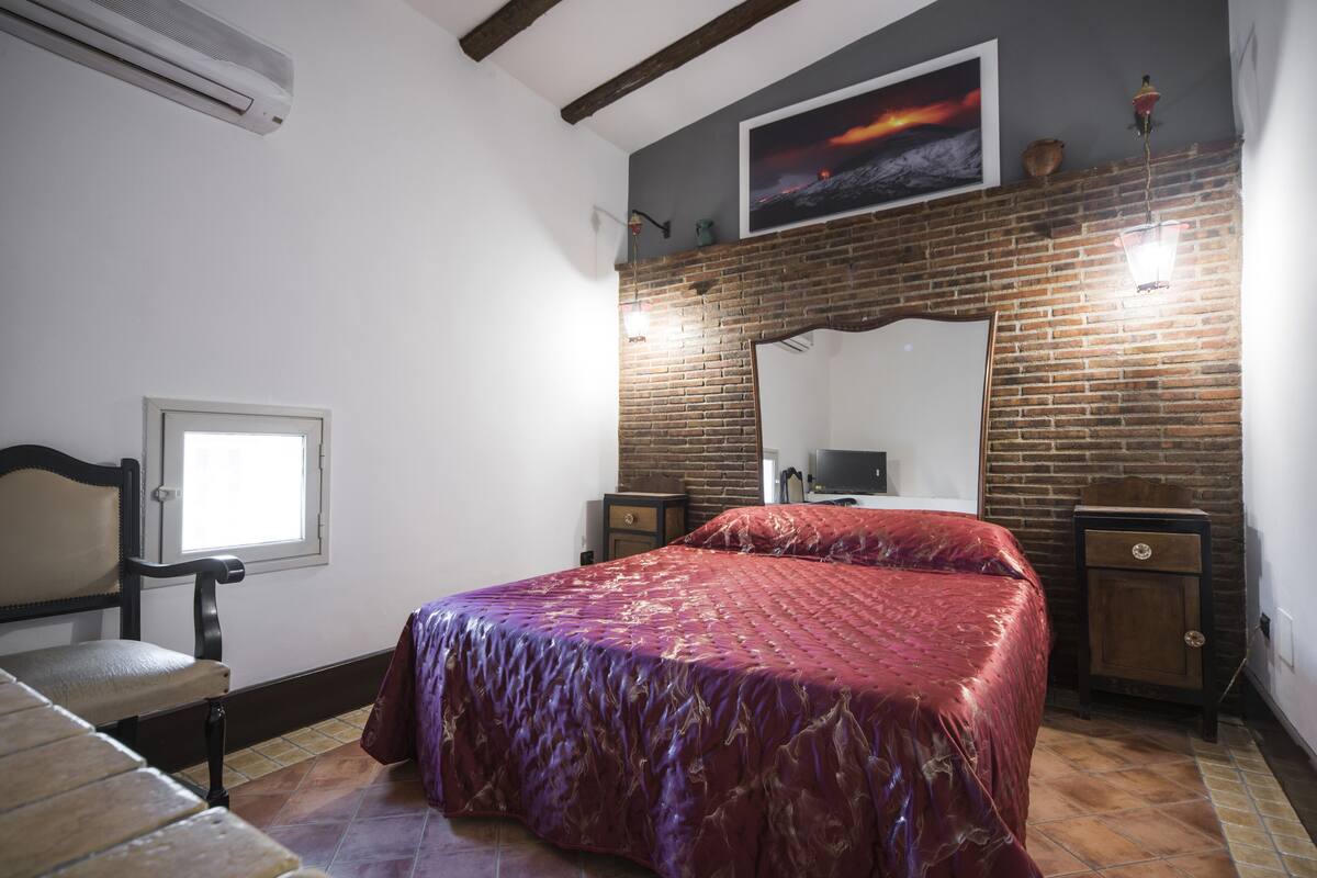 A bedroom with a large bed, a window, a mirror, and a brick wall.