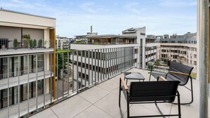 Un balcón moderno con vistas a edificios circundantes, barandilla de vidrio y zona de estar con mesa y sillas.