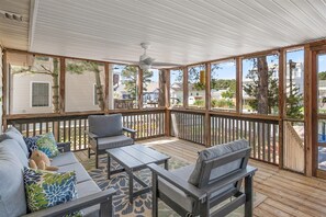 A covered patio with a wooden deck, a gray sofa with patterned cushions, and a ceiling fan.