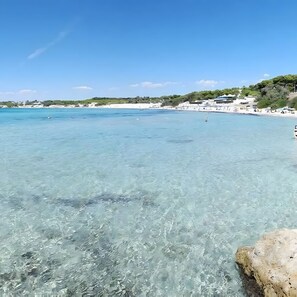 Escena costera con aguas turquesas cristalinas, una playa arenosa y edificios al fondo.