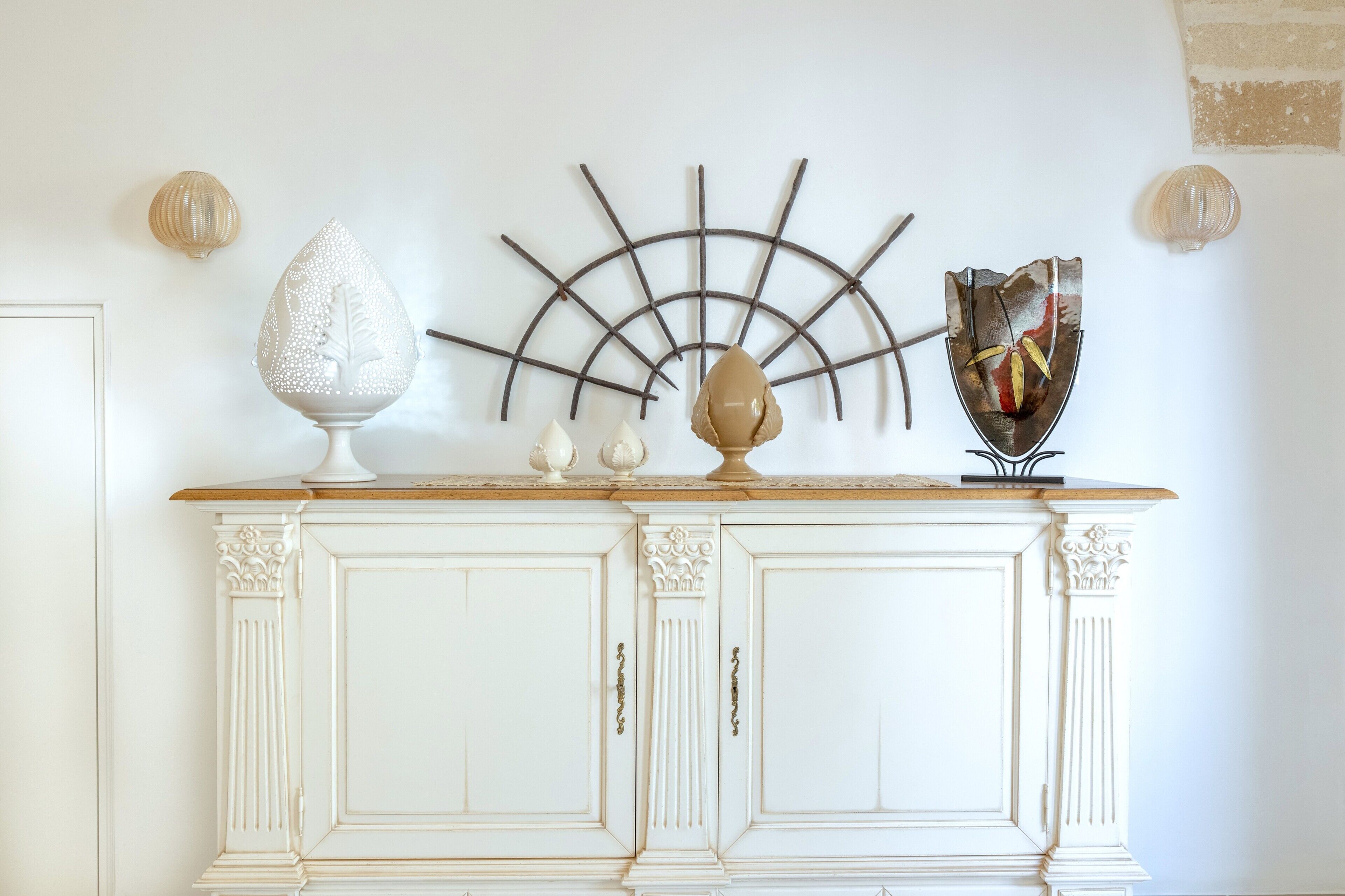 A white cabinet with decorative moldings, a wall-mounted sunburst light, and a few small vases on top.