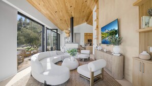 Ein modernes Wohnzimmer mit einer großen L-förmigen Couchgarnitur, einem Couchtisch und einem Fernseher. Es gibt eine Holzbalkendecke, ein großes Fenster und eine Küche im Hintergrund.