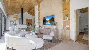 Ein modernes Wohnzimmer mit Kamin, weißen Sofas, einem Flachbildfernseher, Holzregalen und einer Küche im Hintergrund.