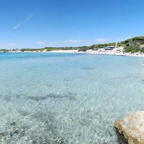 Un paysage côtier avec une eau turquoise et limpide, une plage de sable et des bâtiments en arrière-plan.
