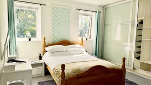 A bedroom with a wooden bed, a desk, and a chair. There are two windows with curtains, a wardrobe, and a small plant on the windowsill.