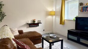 A living room with a brown leather sofa, a black coffee table, a TV stand, a bookshelf, and a window with yellow curtains.