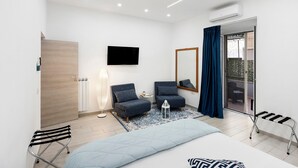 A modern hotel room with a bed, two armchairs, a TV, a mirror, a radiator, and a sliding glass door to the balcony.