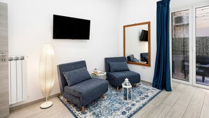 A modern living room with a flat-screen TV, two blue armchairs, a floor lamp, a mirror, and a view through a glass door.