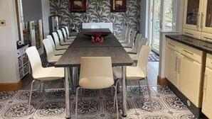 A dining area with a long table, white chairs, patterned floor tiles, and a kitchen with cabinets and a window. - Relaxing 5-star Farm Stay, Sleeps 20 With Spa! (Shotts)