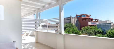 A balcony with a white railing, a wicker sofa, and a view of buildings and greenery.