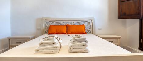 Una cama bien hecha con dos pilas de toallas dobladas, un cabecero blanco y dos mesitas de noche con almohadas naranjas.