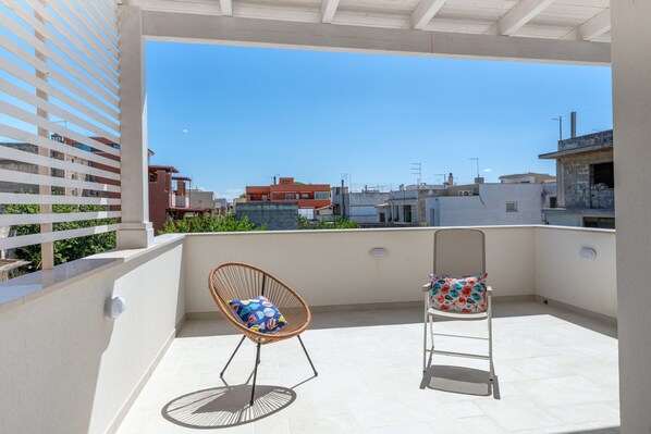 A balcony with two chairs, one with a patterned cushion and another with a floral cushion, overlooking a cityscape with buildings and a clear sky. - Apartment 'Primo Piano - Mini' with Private Garden, Wi-Fi and Air Conditioning (Pulsano)
