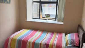 Una habitación pequeña con una cama individual, una ventana con vistas a edificios y un florero con flores en el alféizar.