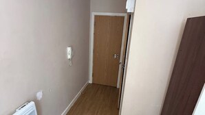 Un pasillo estrecho con una puerta de madera al final, un radiador blanco a la izquierda y una pared blanca con una pequeña mancha de desperfecto.