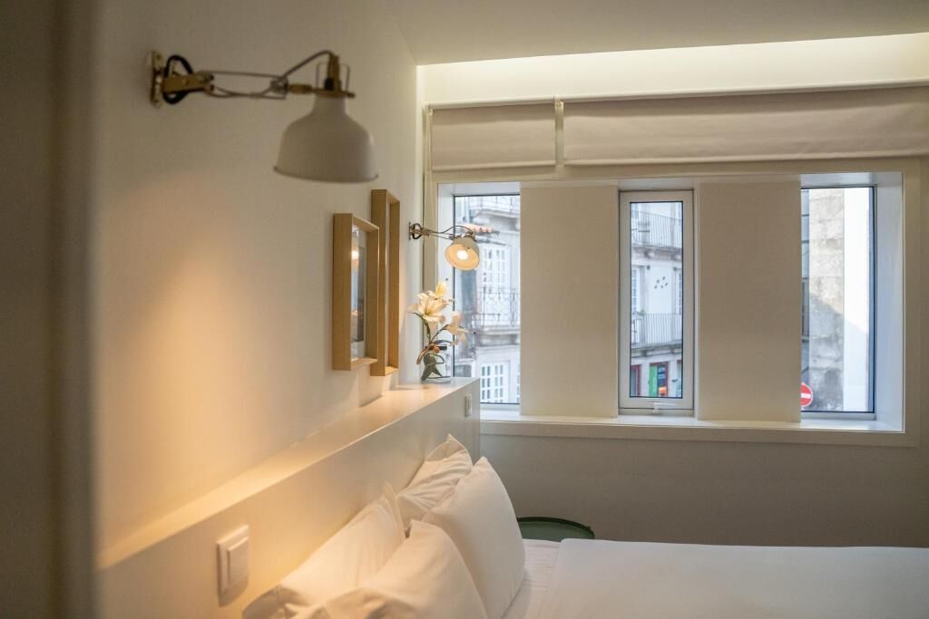 Een hotelkamer met een bed, nachtkastje, spiegel en raam met uitzicht op gebouwen.