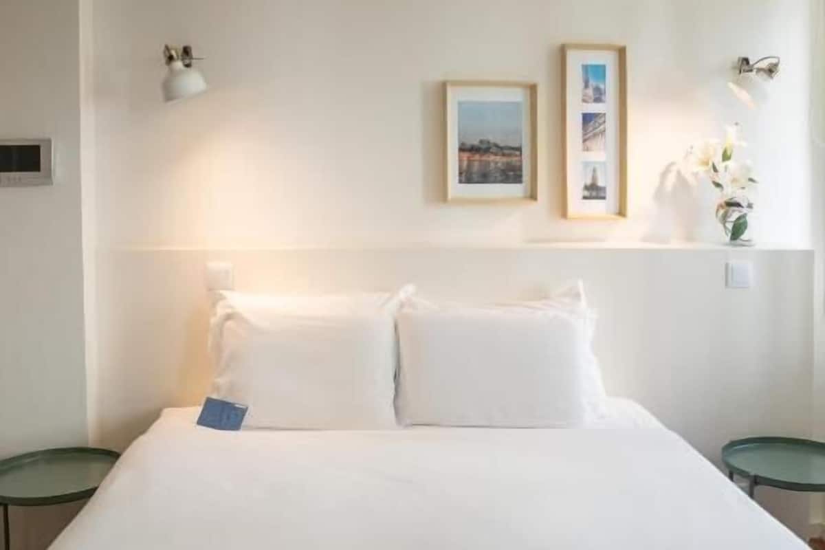 A neatly made bed with white linens, a small bedside table, and a wall-mounted lamp. There are two framed pictures on the wall above the bed.