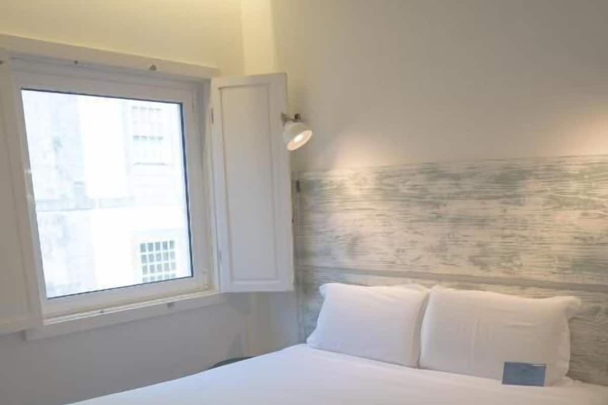A neatly made bed in a room with a window, wooden flooring, and a wall with a textured pattern.