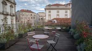 An outdoor patio with tables and chairs, surrounded by plants and overlooking a cityscape with historic buildings. - Oporto Riverside Rooftop (Porto)