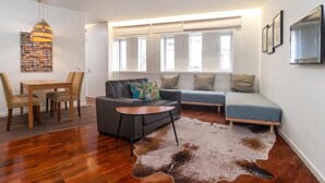 A modern living room with a sofa, a coffee table, and a dining area with chairs. There is a large window with blinds, a hanging light fixture, and a mounted television. - Oporto Riverside Rooftop (Porto)