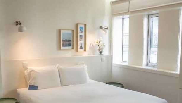 A bedroom with a large bed, wooden flooring, a bedside table, a window with blinds, and wall-mounted lights. - Oporto Riverside Rooftop (Porto)