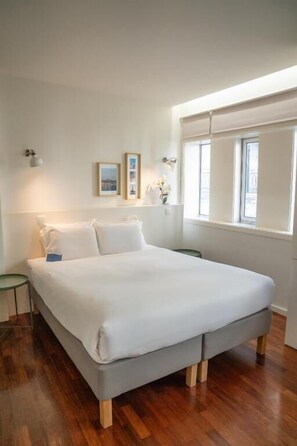 A bedroom with a large bed, wooden flooring, a bedside table, a window with blinds, and wall-mounted lights. - Oporto Riverside Rooftop (Porto)