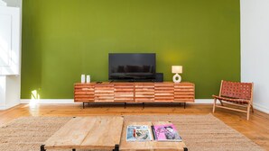 Una sala de estar con pared verde, mueble de entretenimiento de madera, televisor, lámpara, sillón y mesa de centro con revistas.