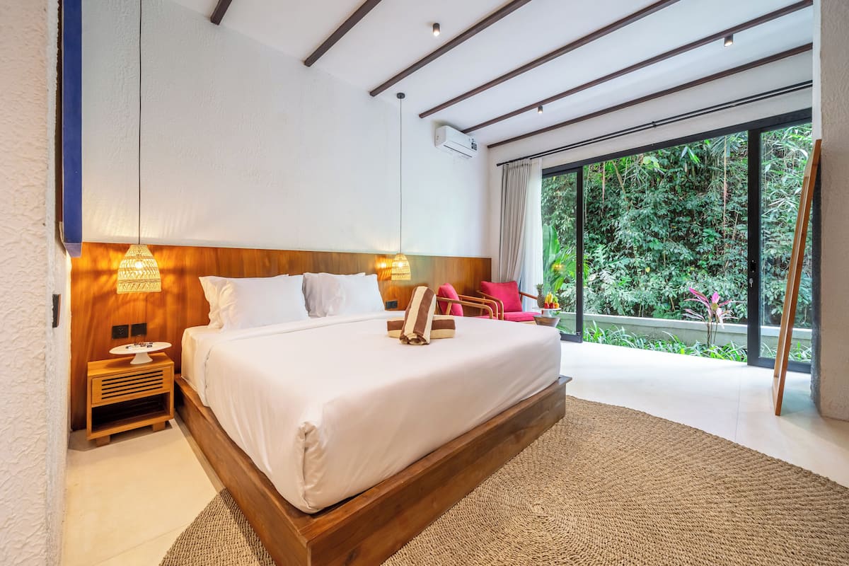 Une chambre moderne avec un grand lit, une tête de lit en bois, des lampes de chevet et une vue sur une végétation luxuriante grâce à de grandes fenêtres.