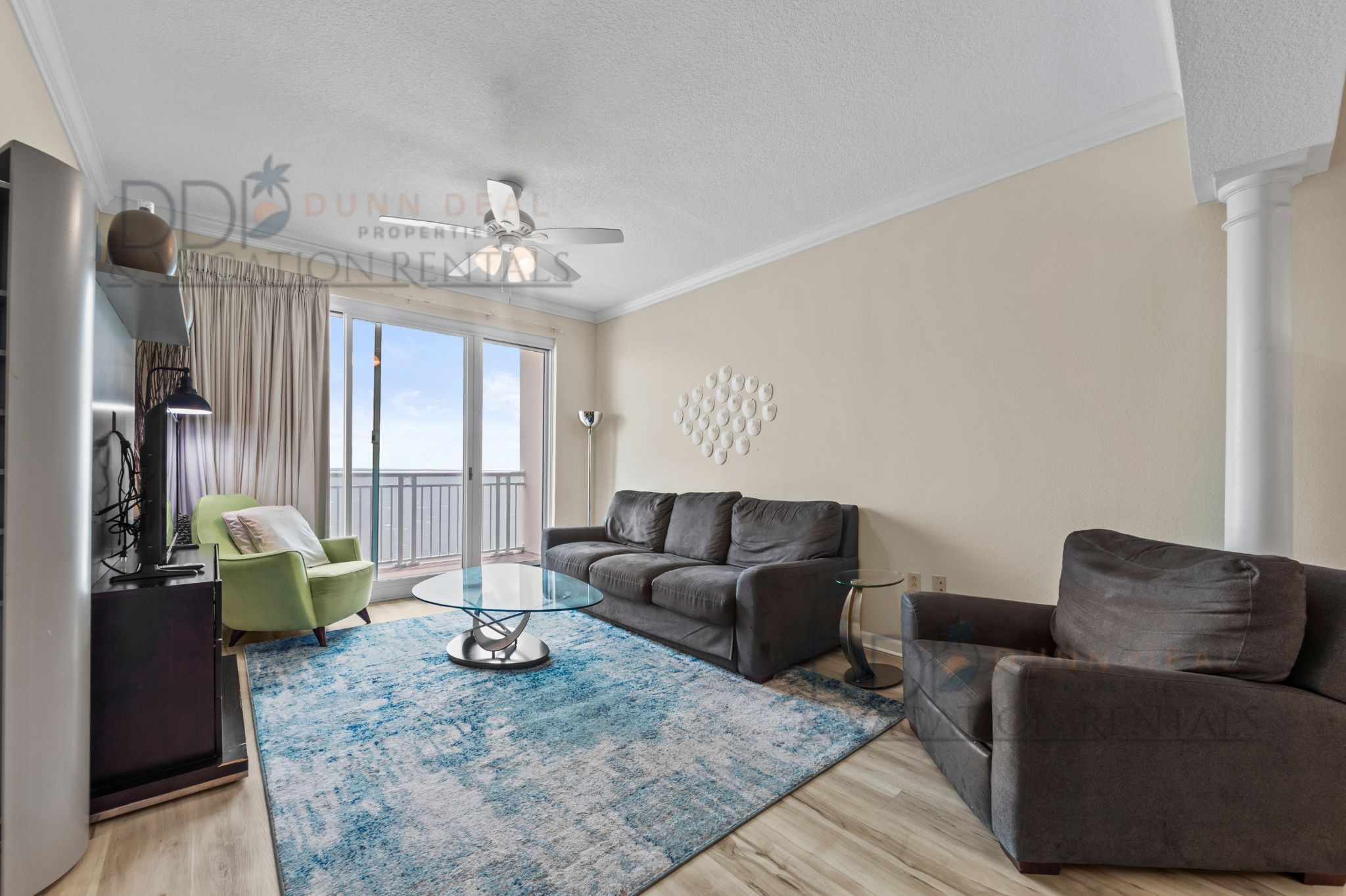 Een moderne woonkamer met een grijze bank, een glazen salontafel, een groene fauteuil en een blauw vloerkleed. Er is een plafondventilator, een groot raam met gordijnen en een decoratief wandobject.