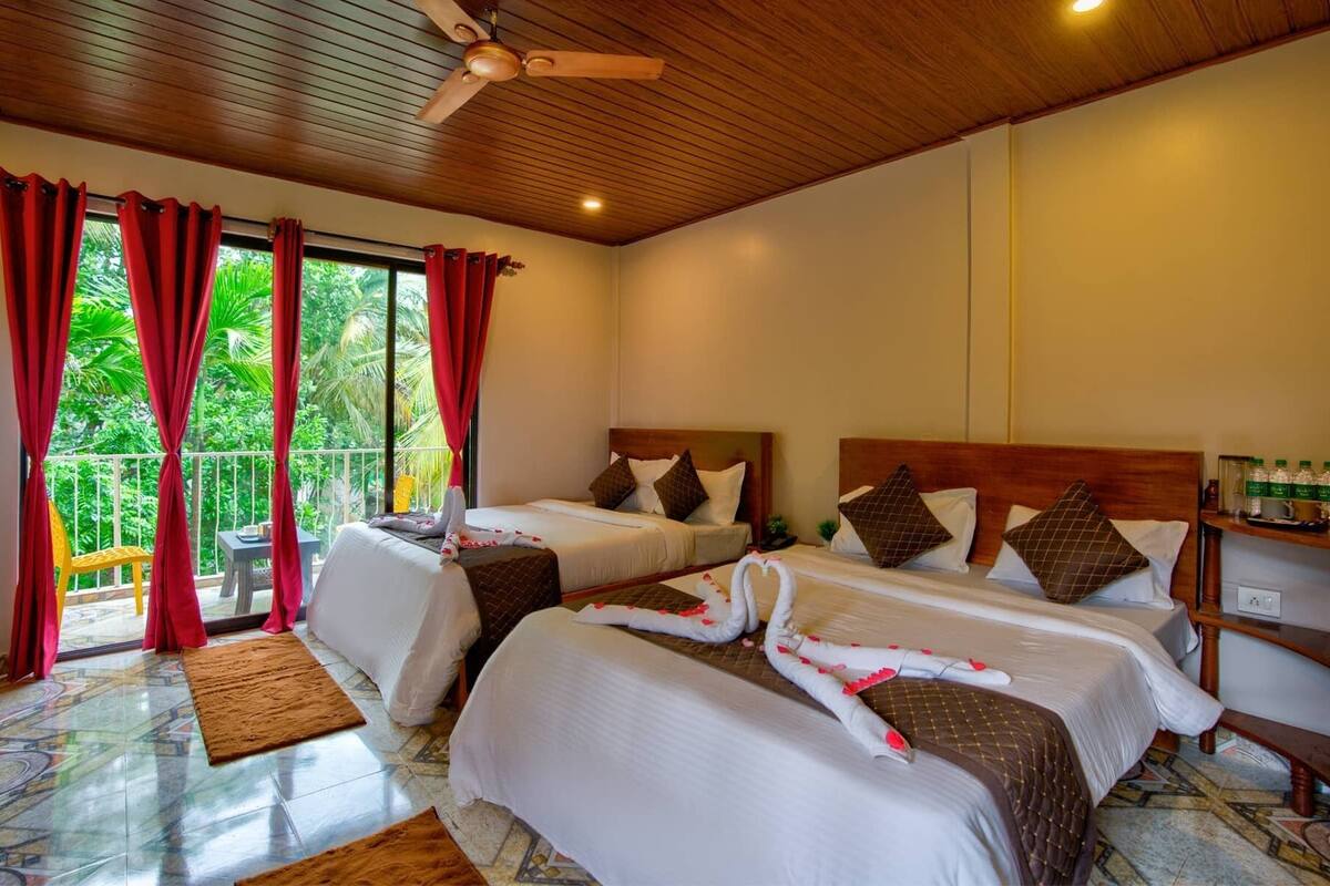 A hotel room with two beds, a balcony with red curtains, a ceiling fan, and a view of greenery.
