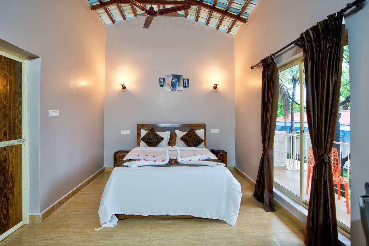 A bedroom with a wooden headboard, a bed with white bedding, two wall-mounted lights, and a view of the outdoors through a window with curtains.