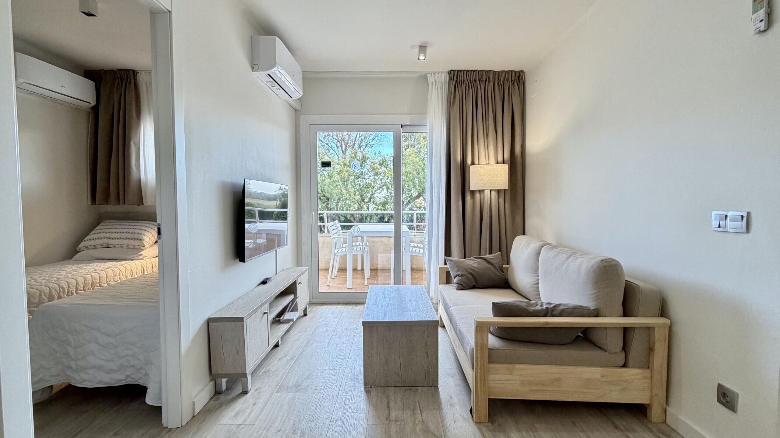 A modern, well-lit room with a bed, a sofa, a TV, and a balcony with a table and chairs.
