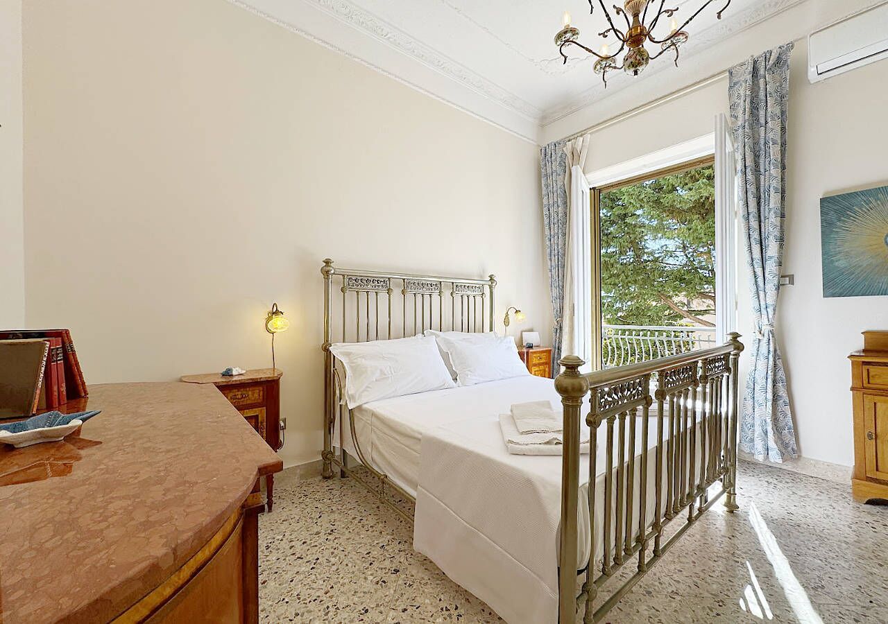 A bedroom with a bed, bedside table, lamp, and a view of the outdoors.