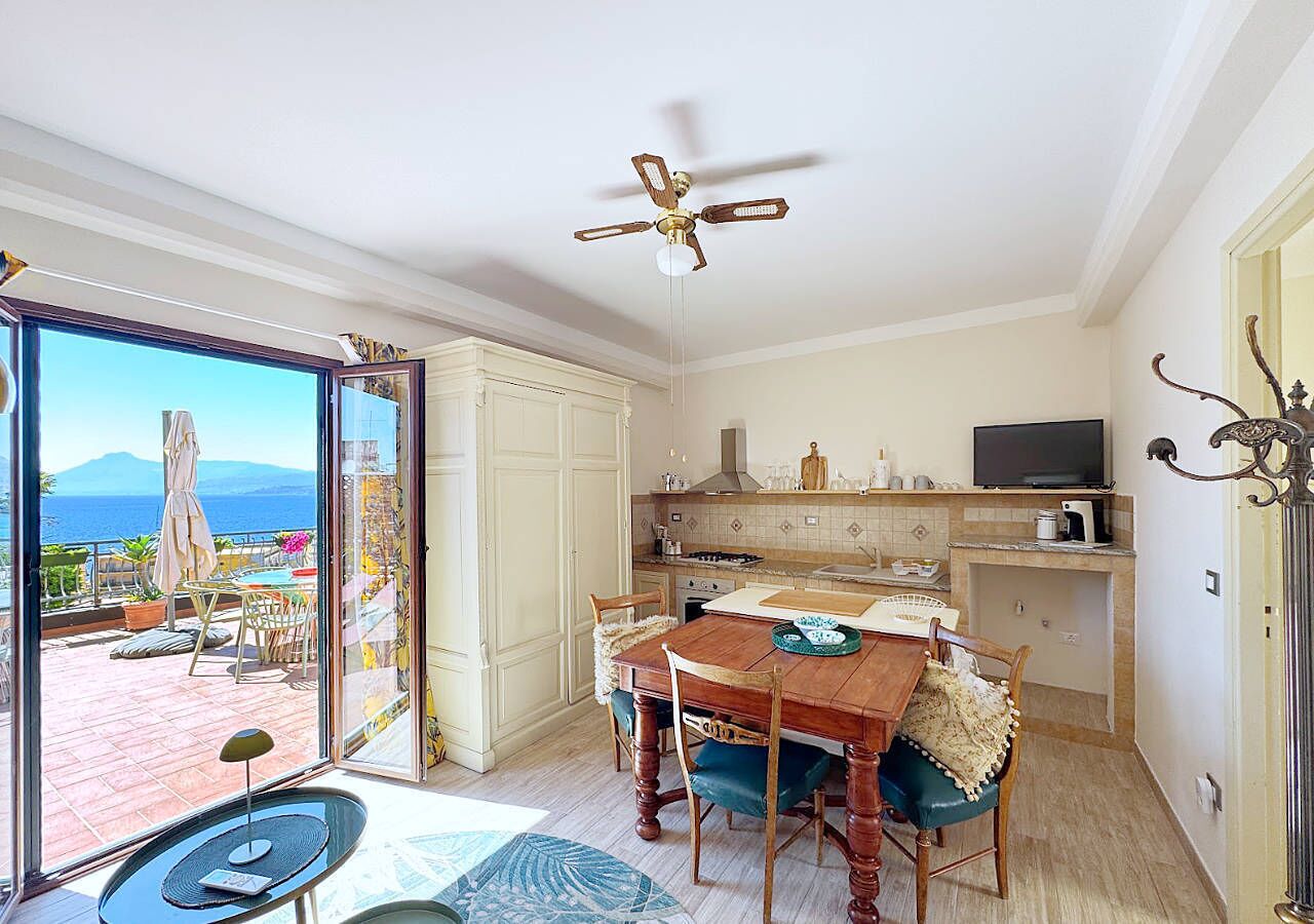 A spacious dining area with a wooden table and chairs, a ceiling fan, and a view of the sea through large glass doors.