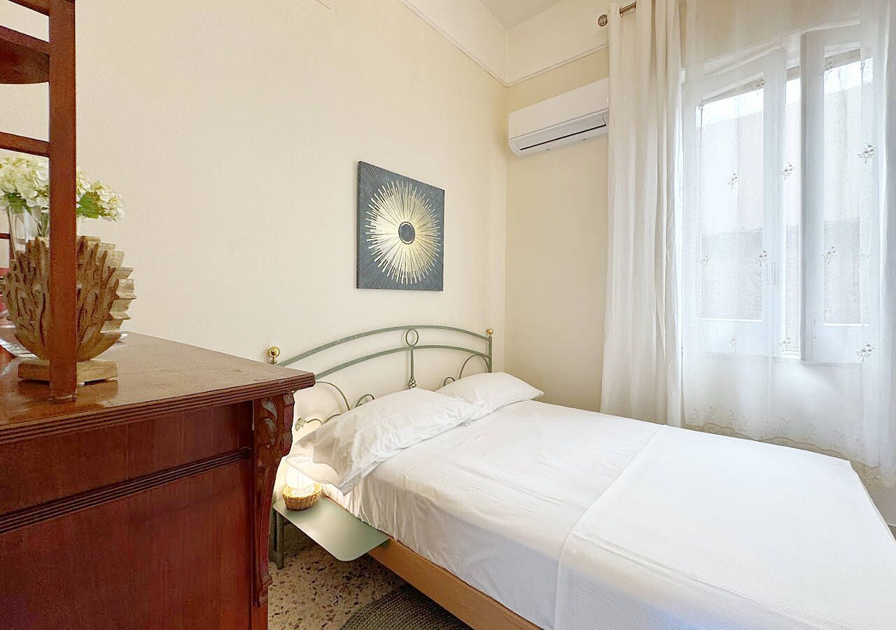 Een slaapkamer met een bed, een houten dressoir, een wandairconditioner en een raam met dunne gordijnen.