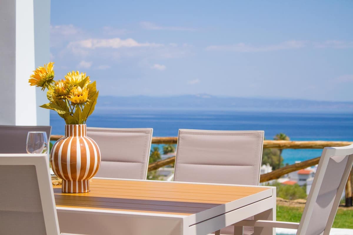 A table with a striped vase and chairs, overlooking a coastal view.