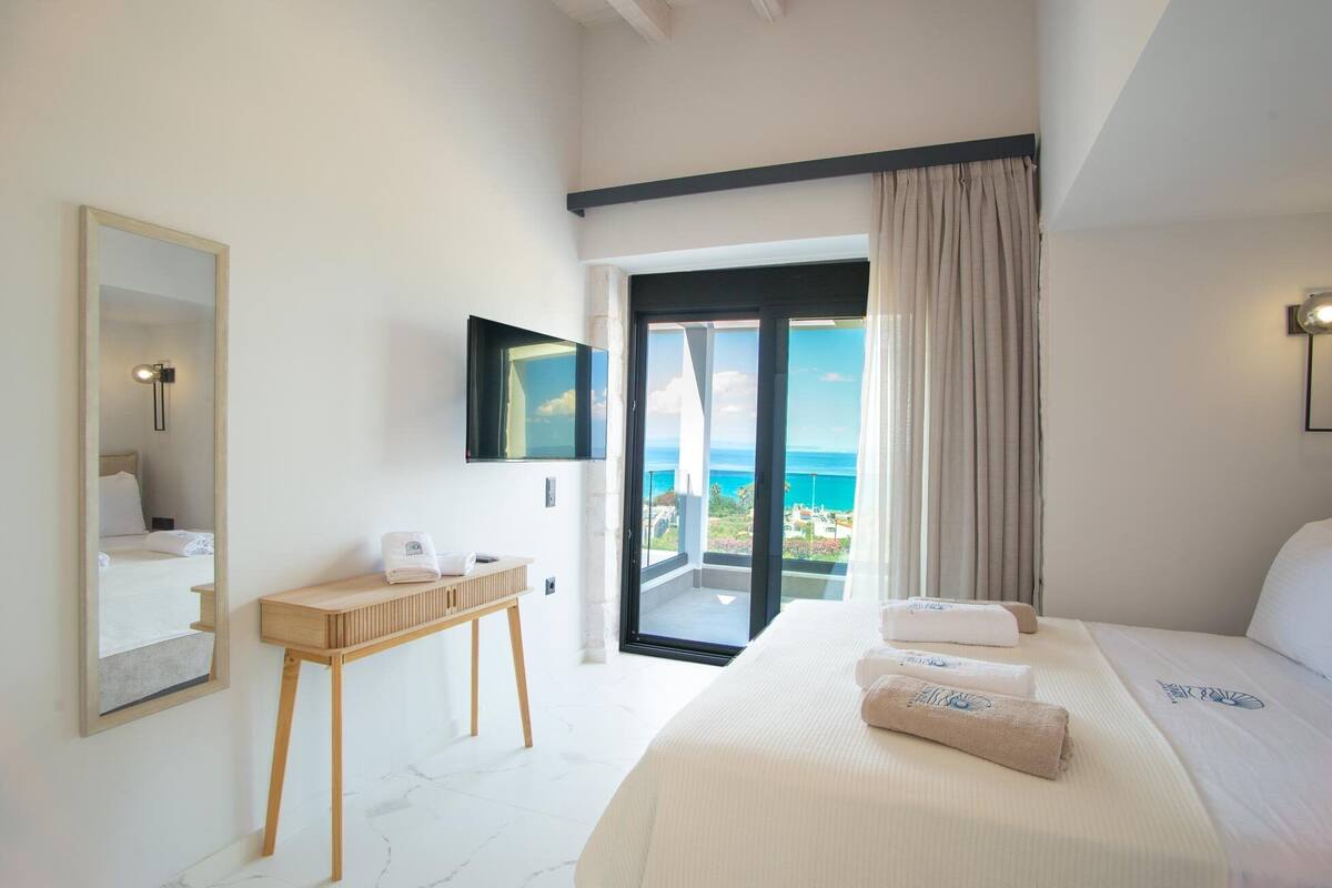 A hotel room with a bed, a bedside table, a mirror, a TV, and a view of the ocean.