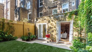 A small backyard with a wooden fence, a barbecue grill, and a door leading to a room with a bed and a table. - Brunswick Gardens II by Onefinestay (London)