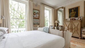 A spacious bedroom with a large bed, a dresser, and a mirror. There are windows with curtains, a painting on the wall, and a lamp on the dresser. - Brunswick Gardens II by Onefinestay (London)