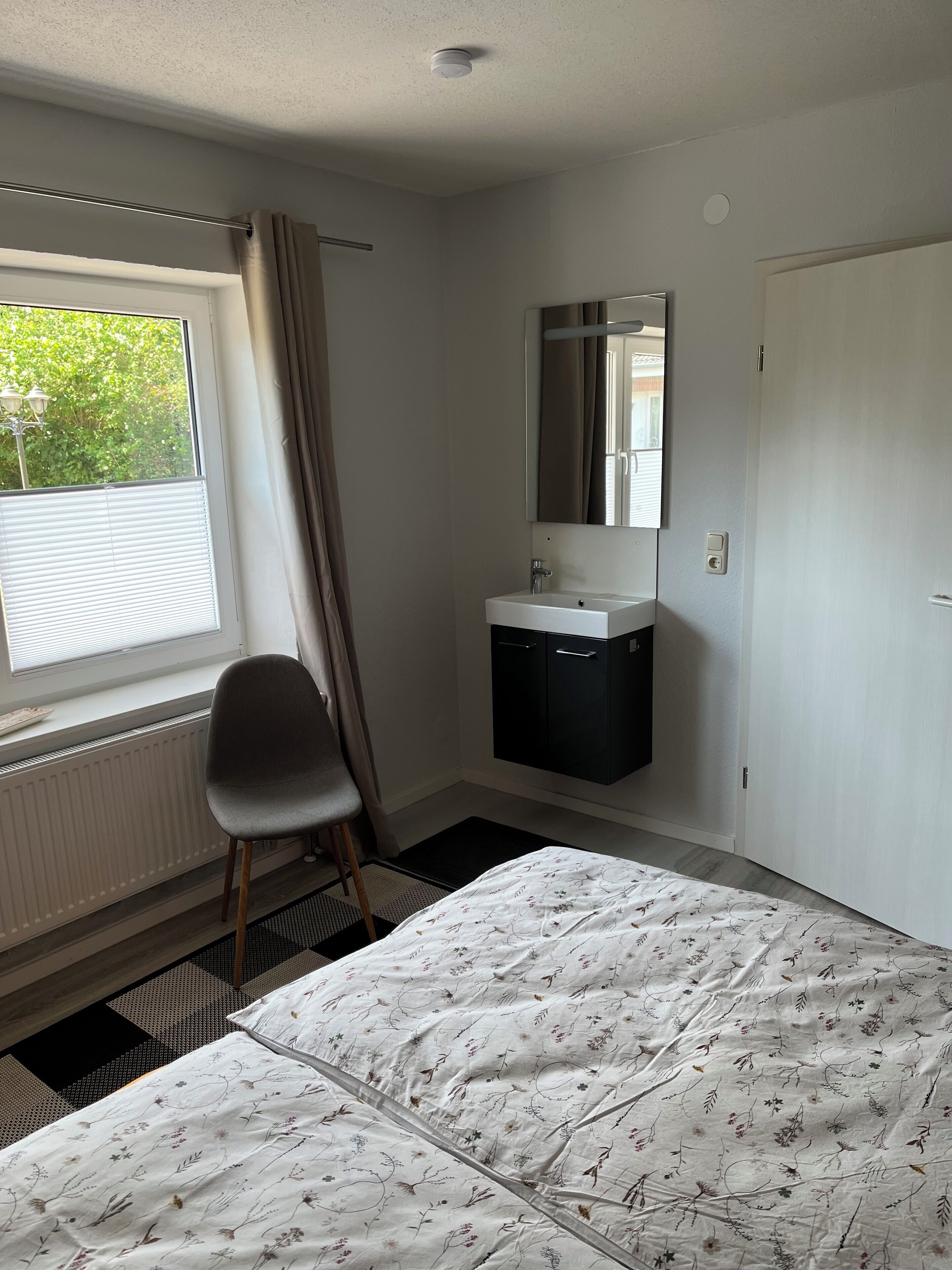 A bedroom with a bed, a chair, a window with curtains, a radiator, a sink, and a mirror.