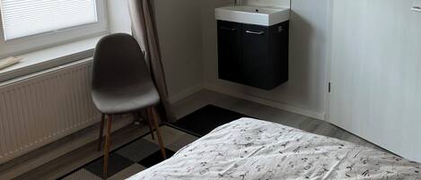 A bedroom with a bed, a chair, a window with curtains, a radiator, a sink, and a mirror.