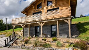 A modern wooden house with a balcony, stone foundation, and a grassy landscape.