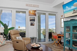 A living room with a beige sofa, a coffee table with seashells, a teal cabinet with decorative items, and a TV displaying a beach scene.